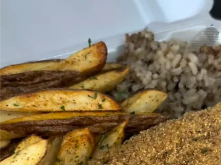 Seasoned potato wedge fries and rice beside a big cornmeal-fried fish fillet in a takeout box